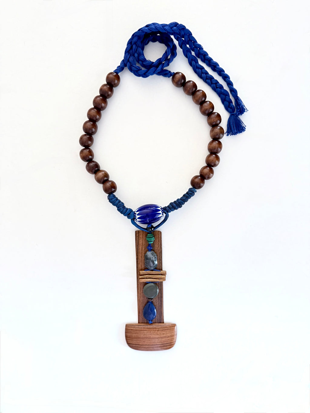  A quiet sculptural presence. One-of-a-kind sculptural necklace built around an  architectural bookmark wooden pendant — vertical, solid, grounded. Various stones, golden coral, a Venetian (Murano)chevron trade glass bead, wood beads on Cobalt blue adjustable braided threads complete this unique piece handmade in Lebanon with materials sourced all over the world. 
An Architectural and tactile totem. 